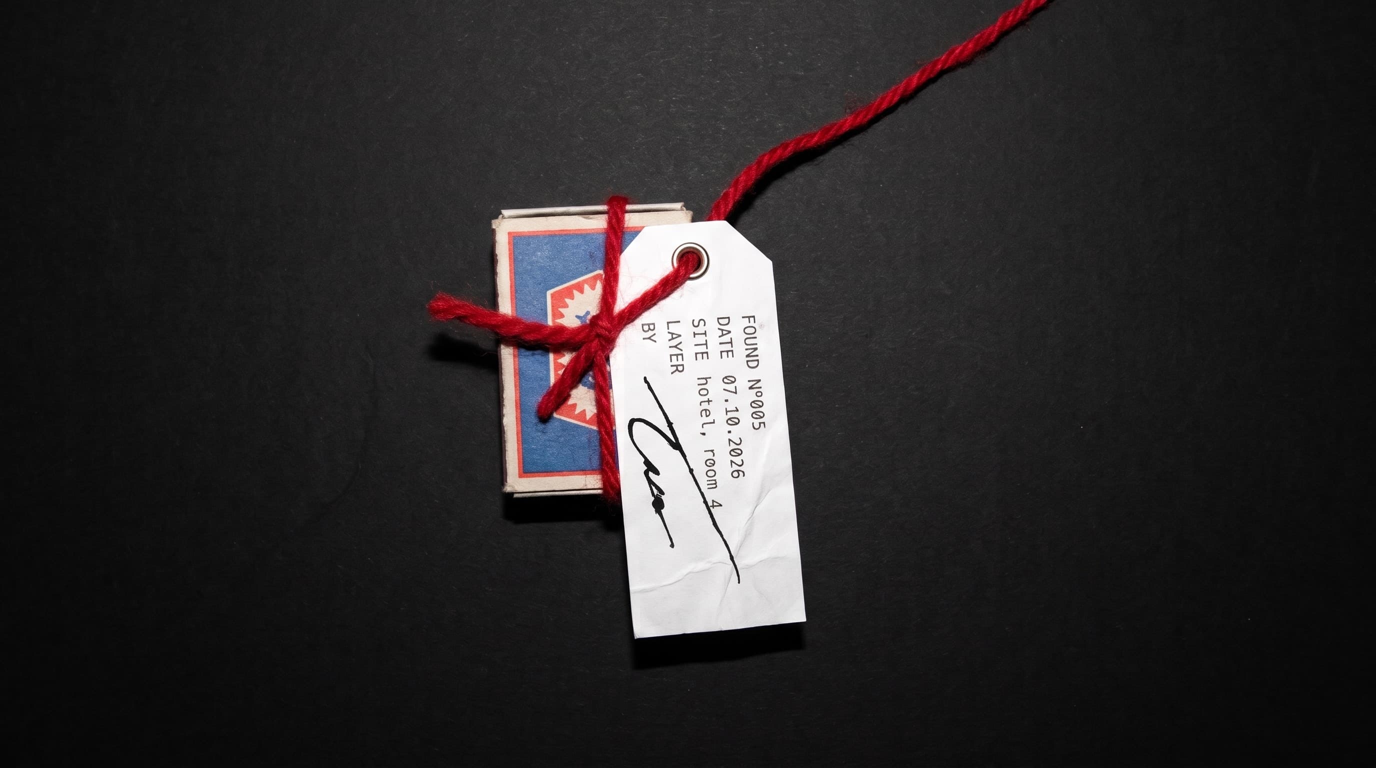 A worn matchbox tied with red wool. A tag on the thread reads Found N°005, 07.10.2026, hotel, room 4, and is signed by hand.